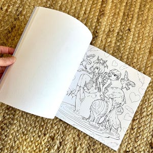 以下が含まれることがあります： 開かれた塗り絵本。詳細な白黒イラストで、鳥とハートを持つ2人のアニメ風の人物が描かれています。本は開かれており、空白のページとアートワークが表示されています。