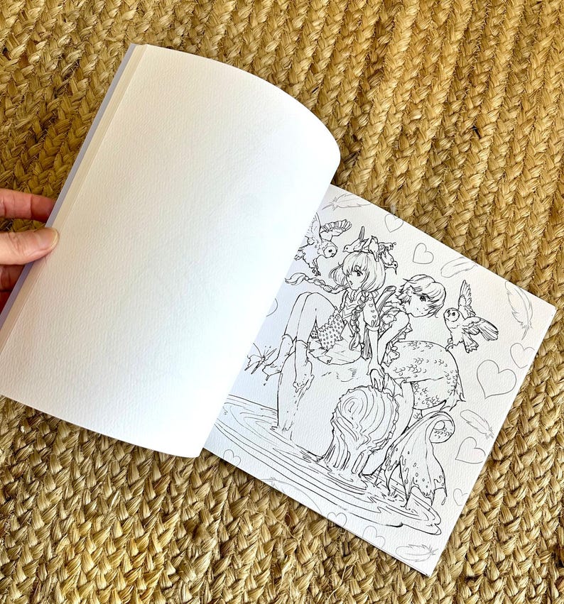 以下が含まれることがあります： 開かれた塗り絵本。詳細な白黒イラストで、鳥とハートを持つ2人のアニメ風の人物が描かれています。本は開かれており、空白のページとアートワークが表示されています。