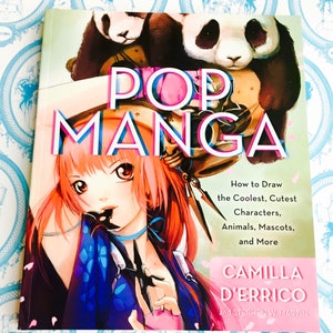 Peut inclure: Un livre intitulé "POP MANGA" avec une illustration manga d'un personnage aux cheveux roses et d'un panda. La couverture comprend également le texte "How to Draw the Coolest, Cutest Characters, Animals, Mascots, and More." Le livre est de Camilla D'Errico et Stephen W. Martin.