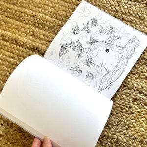 以下が含まれることがあります： 蝶と鳥が描かれた生き物の詳細な白黒イラストが特徴の、開いた塗り絵。ページは白く、本は織られたベージュ色の表面に置かれています。
