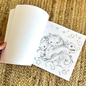 以下が含まれることがあります： 3匹のドラゴンが描かれた、白黒のイラストが特徴的な塗り絵が開かれています。ドラゴンはとげのある特徴があり、雲のような形に囲まれています。本は織られたベージュ色の表面に置かれています。