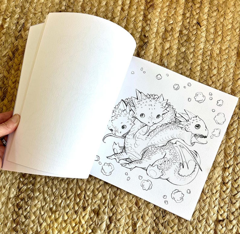 以下が含まれることがあります： 3匹のドラゴンが描かれた、白黒のイラストが特徴的な塗り絵が開かれています。ドラゴンはとげのある特徴があり、雲のような形に囲まれています。本は織られたベージュ色の表面に置かれています。