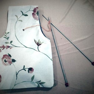 Puede incluir: Una tela blanca con un patrón floral, dos agujas de tejer con puntas rojas y un trozo de hilo naranja.