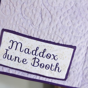 May include: Purple quilted blanket with a white label sewn on. The label has gold glitter and the text "Maddox June Booth" in cursive.