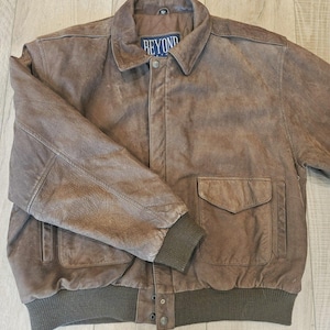 Peut inclure: Un blouson aviateur en cuir marron doté d'un col rabattu, d'une fermeture à boutons sur le devant et de deux grandes poches à rabat. Le blouson présente des poignets côtelés marron foncé et un ourlet côtelé assorti. L'étiquette indique BEYOND.
