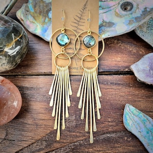 May include: Gold dangle earrings with a labradorite gemstone and a fringe of thin gold bars. The earrings are on a wooden surface with a tag that reads "Song Design, Sonoma County".