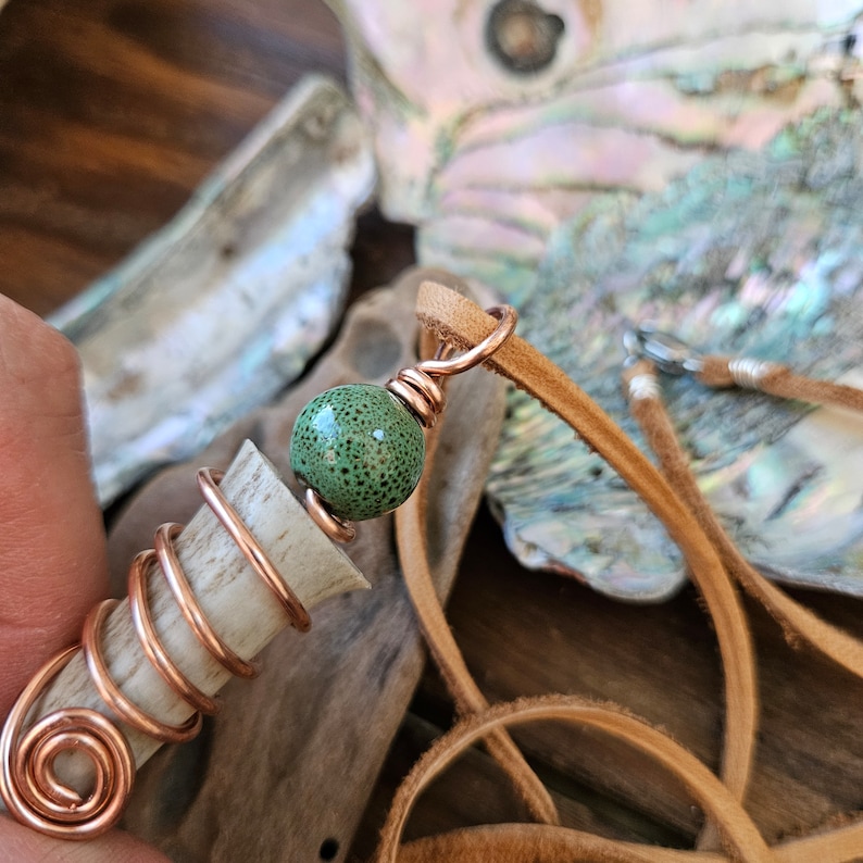 Puede incluir: Una cuenta verde moteada con alambre de cobre envuelto alrededor de un trozo de asta. El asta est&aacute; envuelta en alambre de cobre y unida a un cord&oacute;n de cuero marr&oacute;n.