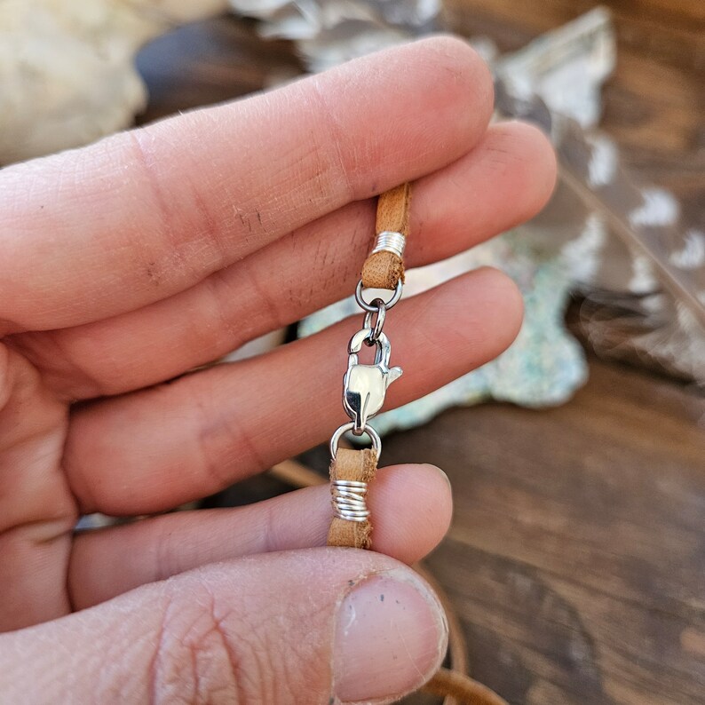 Puede incluir: Primer plano de un collar de cord&oacute;n de cuero marr&oacute;n con un cierre plateado. El cord&oacute;n est&aacute; envuelto con alambre plateado en el cierre.
