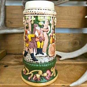 May include: A cylindrical ceramic beer stein featuring a raised relief scene of figures in a forest setting. The stein has a cream-colored top rim, a central band with painted figures in yellow and brown, and a green base with floral motifs.