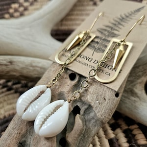 May include: Gold earrings with a cowrie shell charm and a geometric design. The earrings are made with a gold chain and a gold hook. The earrings are made by Song Designs, Sonoma County, CA.