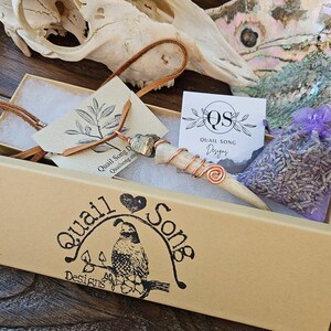Puede incluir: Una caja marr&oacute;n con un logotipo de codorniz y el texto "Quail Song Designs". La caja contiene un collar con un colgante de hueso envuelto en alambre de cobre, una peque&ntilde;a bolsa de lavanda y una tarjeta con el texto "Quail Song Designs".
