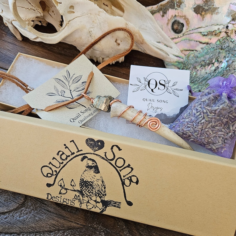 Puede incluir: Una caja marr&oacute;n con un logotipo de codorniz y el texto "Quail Song Designs". La caja contiene un collar con un colgante de hueso envuelto en alambre de cobre, una peque&ntilde;a bolsa de lavanda y una tarjeta con el texto "Quail Song Designs".