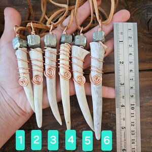 Puede incluir: Seis colgantes de asta de ciervo con envoltura de alambre de cobre y acentos de piedra de pirita. Los colgantes est&aacute;n numerados del 1 al 6 y se muestran sobre un fondo de madera con una regla para referencia de tama&ntilde;o.