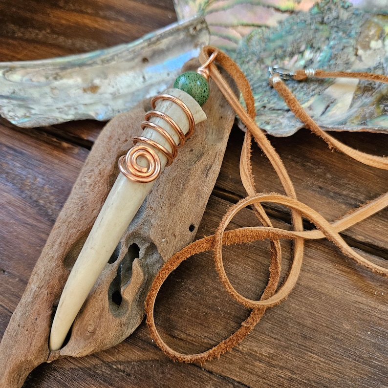 Puede incluir: Un collar con un colgante en forma de hueso envuelto en alambre de cobre y una cuenta de piedra verde. El collar est&aacute; en un cord&oacute;n de cuero marr&oacute;n.