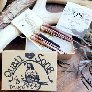 Könnte beinhalten: Ein Paar Creolen mit federartigen Anhängern in Braun-, Rosa- und Weißtönen. Die Ohrringe werden in einer kleinen Schachtel mit dem Logo von Quail Song Designs präsentiert. Die Schachtel besteht aus braunem Karton und hat eine Herzform mit dem aufgedruckten Schriftzug "Quail Song Designs".