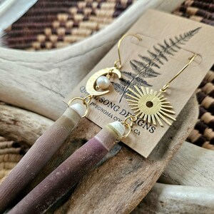 May include: A pair of gold earrings with a crescent moon charm, a pearl, and a sunburst charm. The earrings are made with natural materials and are handmade in Sonoma County, CA.  "Quel Song Designs"