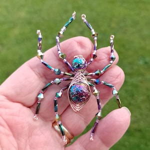 May include: A handcrafted, iridescent spider sculpture. The spider's body is made of a wire-wrapped, multi-colored glass bead. The legs are constructed of iridescent beads and wire, with the legs and body in shades of purple, green, and blue. The spider is held in a hand.