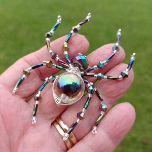 以下が含まれることがあります： 虹色のビーズと大きな輝くオーブボディを備えた手作りのクモの彫刻。脚は、緑、青、紫の色合いのファセットビーズでできており、銀色のワイヤーで接続されています。クモは手で持たれています。