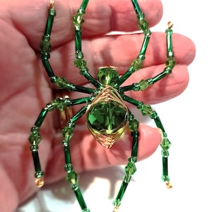 Op de afbeelding: Een handgemaakt spinnenornament met een groene edelsteenlichaam en poten van groene kralen en draad. Het lichaam van de spin is omwikkeld met goudkleurig draad. De spin wordt in de hand van een persoon tegen een witte achtergrond gehouden.