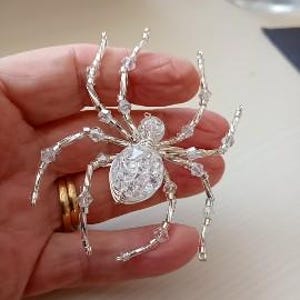 Puede incluir: Broche de araña plateado con detalles de cristal transparente. El cuerpo de la araña es un grupo de cristales, y sus patas son de alambre con cuentas de cristal. El broche se sostiene en una mano.