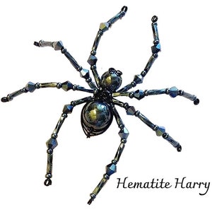 May include: A beaded spider ornament in dark grey. The spider has a rounded body and eight articulated legs. The text "Hematite Harry" is written in a cursive font near the bottom right corner.