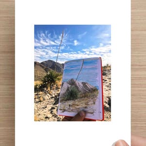 May include: A hand holds a sketchbook with a drawing of a desert landscape. The drawing shows a mountain range, a blue sky with clouds, and a few small plants. The sketchbook is open to the drawing, and the hand is holding it up to the camera. The background of the image is a real desert landscape with a blue sky and clouds.