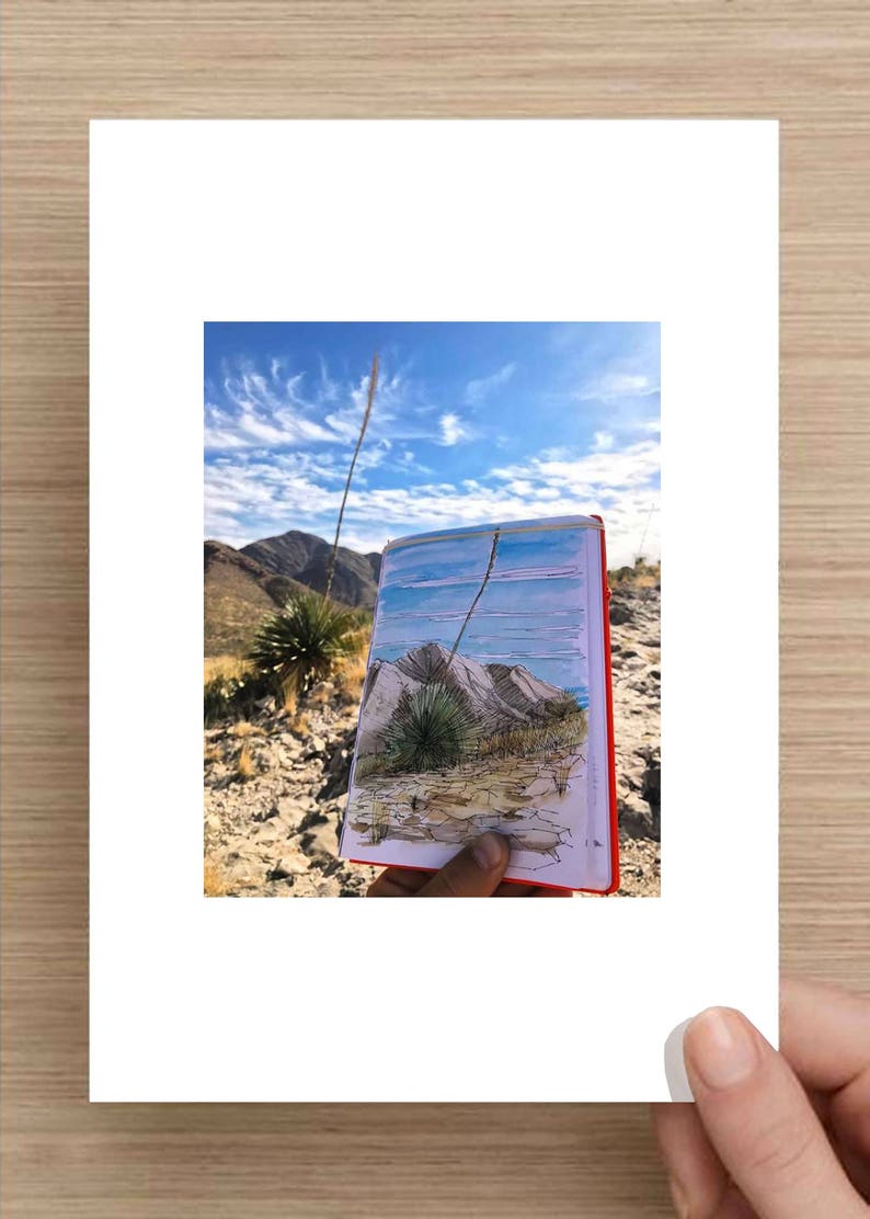 May include: A hand holds a sketchbook with a drawing of a desert landscape. The drawing shows a mountain range, a blue sky with clouds, and a few small plants. The sketchbook is open to the drawing, and the hand is holding it up to the camera. The background of the image is a real desert landscape with a blue sky and clouds.