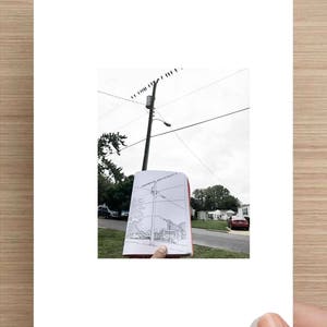May include: A person holds a sketchbook with a drawing of a utility pole with power lines and a street scene. The drawing is being compared to the actual scene in the background.