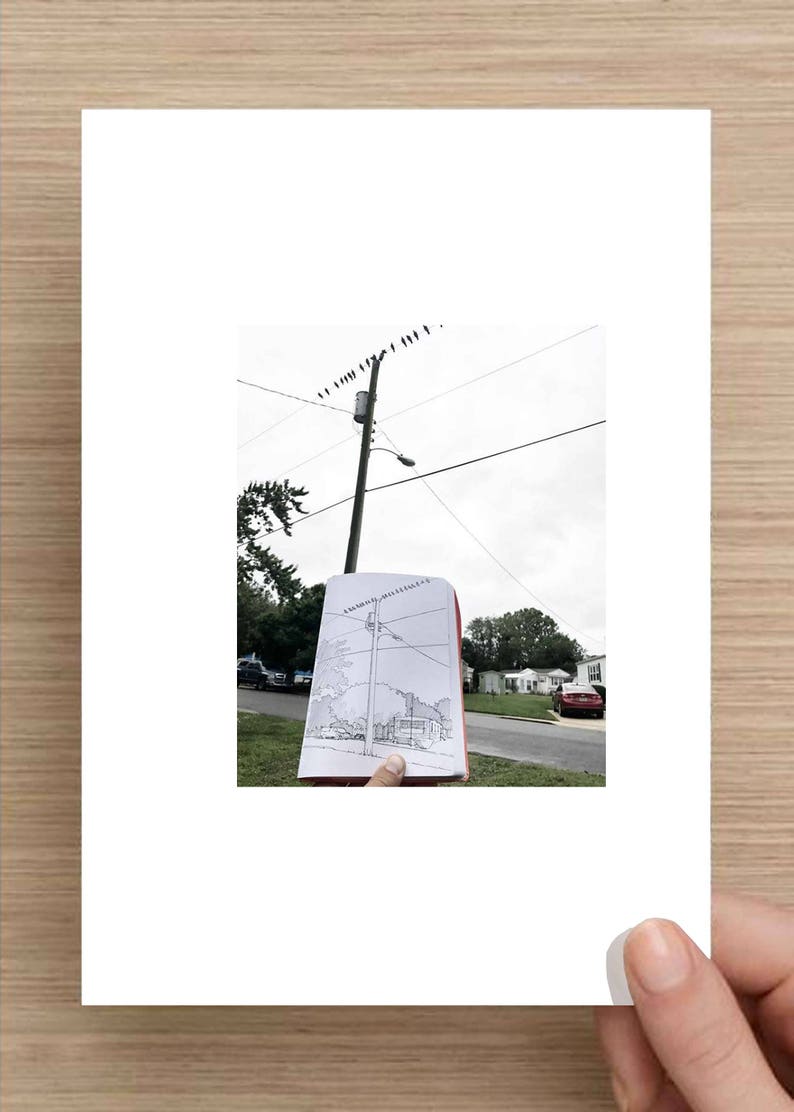 May include: A person holds a sketchbook with a drawing of a utility pole with power lines and a street scene. The drawing is being compared to the actual scene in the background.