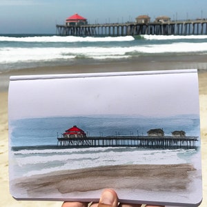 May include: A watercolor painting of a pier with a red roof extending over the ocean. The painting is on a white piece of paper and is being held by a hand.