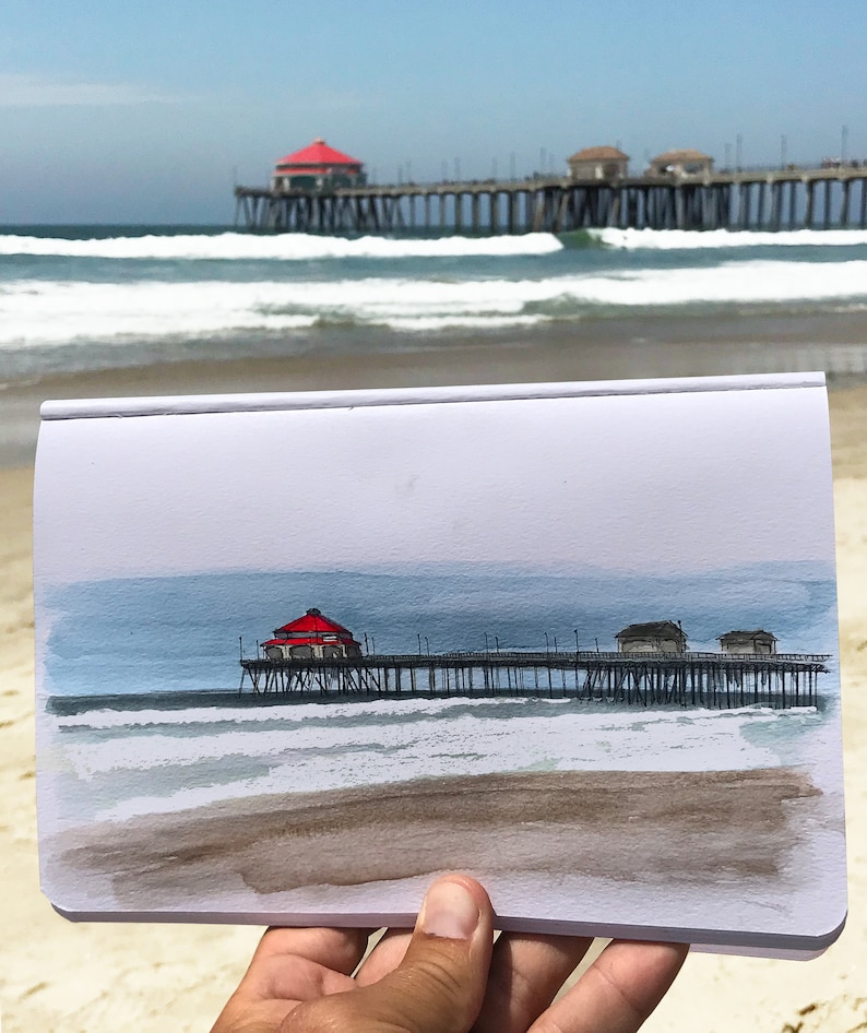 May include: A watercolor painting of a pier with a red roof extending over the ocean. The painting is on a white piece of paper and is being held by a hand.