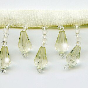 May include: A close-up of a white beaded trim with teardrop-shaped beads. The beads are clear and have a slight iridescent sheen.