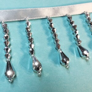 May include: Silver beaded trim with teardrop-shaped beads hanging from a white ribbon.