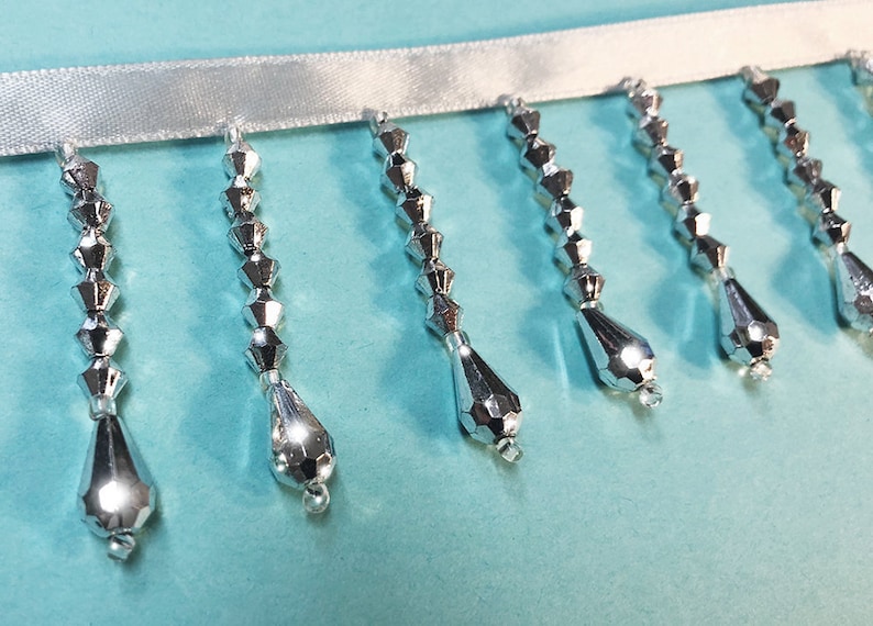 May include: Silver beaded trim with teardrop-shaped beads hanging from a white ribbon.