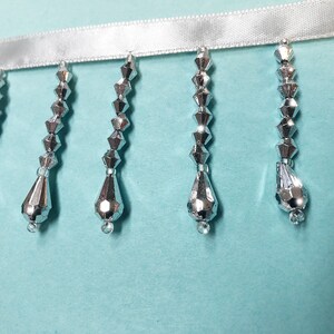 May include: Silver beaded fringe trim with a white satin ribbon backing. The fringe features teardrop-shaped beads.
