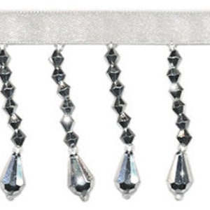 May include: Silver beaded fringe trim with teardrop-shaped beads. The fringe is attached to a white ribbon.