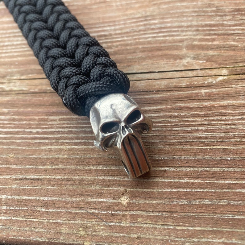 May include: A black paracord bracelet with a silver skull charm. The skull has a detailed design with a prominent jawline.