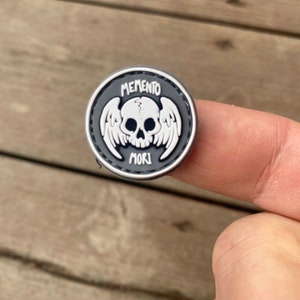 以下が含まれることがあります： 翼のある頭蓋骨と「Memento Mori」というテキストが書かれた白黒のパッチです。