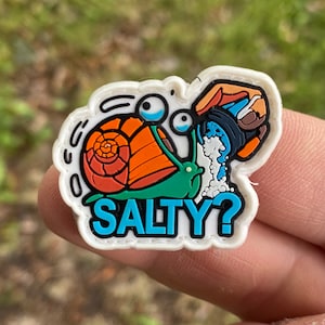 Peut inclure: Un charm de chaussure coloré représentant un escargot de dessin animé avec une coquille orange et un corps vert. L'escargot a de grands yeux expressifs et est arrosé d'eau. Le mot "SALTY?" est écrit en bleu.