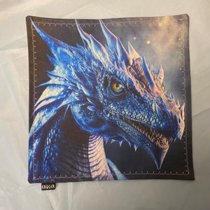 May include: A decorative pillow featuring a blue dragon with a fierce expression and a yellow eye. The pillow has a dark blue background and a white border with a stitched design.