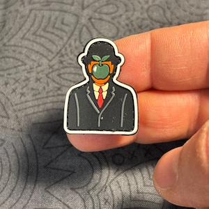 May include: A black and white patch depicting a man in a suit with an apple for a face. The apple is green with orange accents and a red tie. A distinctive accessory.