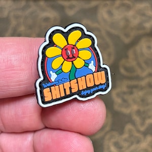 May include: A black and white patch featuring a yellow flower with a red face and blue eyes. The flower has a green stem and is encircled by a blue circle. The text "Welcome to the SHITSOW Enjoy your stay!" is written in orange and white lettering.