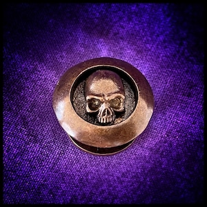 Pode incluir: Um botão redondo de metal de cor bronze com um design de caveira em relevo. A caveira é detalhada com órbitas oculares, um nariz e uma boca.