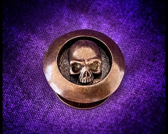 May include: A round, bronze-colored metal button with a raised skull design. The skull is detailed with eye sockets, a nose, and a mouth.