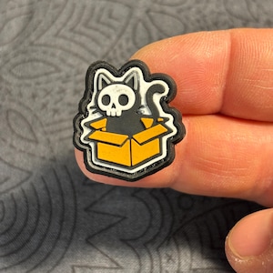 May include: A black and white cat skull design in a yellow box. The cat is sitting in a box with a black outline. The cat has a skull face. The box is yellow with a black outline. The design is on a black background.