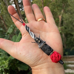 May include: A black and white braided cord with a silver skull charm and a red knot at the end. The cord is attached to a small loop.