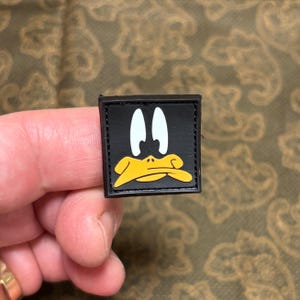 May include: A square, black patch featuring a cartoon duck face with white eyes and a yellow beak. The patch is likely made of fabric or a similar material, with a stitched border. The design is a recognizable cartoon character.