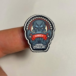 Op de afbeelding: Een zwarte en rode rubberen patch met een blauwe en witte illustratie van een monster met de tekst "OBEY VOTE CTHULHU".