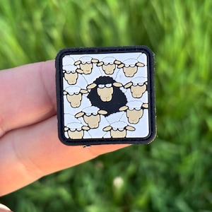 May include: Square patch featuring a black sheep surrounded by white sheep with beige faces. The sheep are arranged on a white background with a black border. The patch is held against a blurred green background.