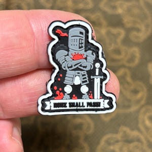 May include: A black and white patch with a cartoon knight in armor. The knight is standing with his arms crossed and a sword at his side. The patch has a red splash of color on the knight's chest and a white banner with the text "None Shall Pass".